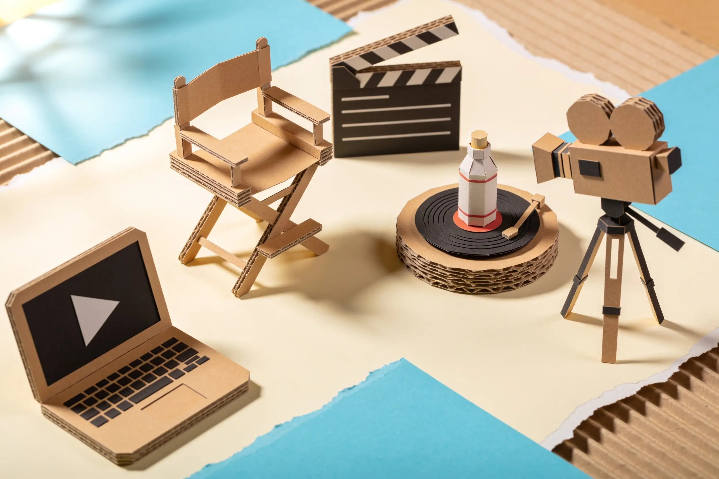 Papercraft film set: a cardboard camera on a tripod pointed at a small product bottle on a turntable, a clapperboard and a paper laptop showing a play button